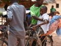Een man helpt een jonger met konzo achterop de fiets            