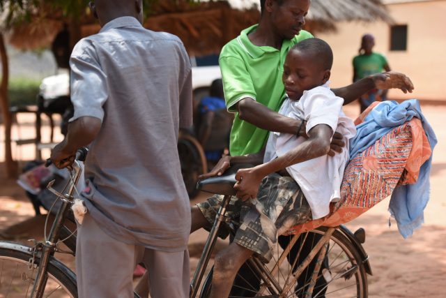 Een man helpt een jonger met konzo achterop de fiets